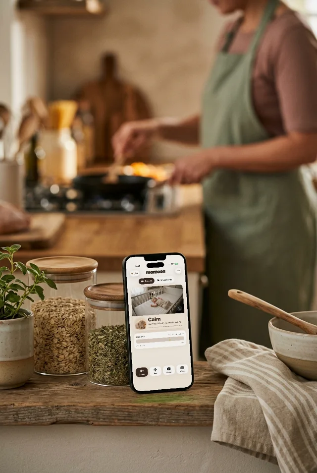 A phone with Mamoon on a kitchen shelf while a parent cooks behind it